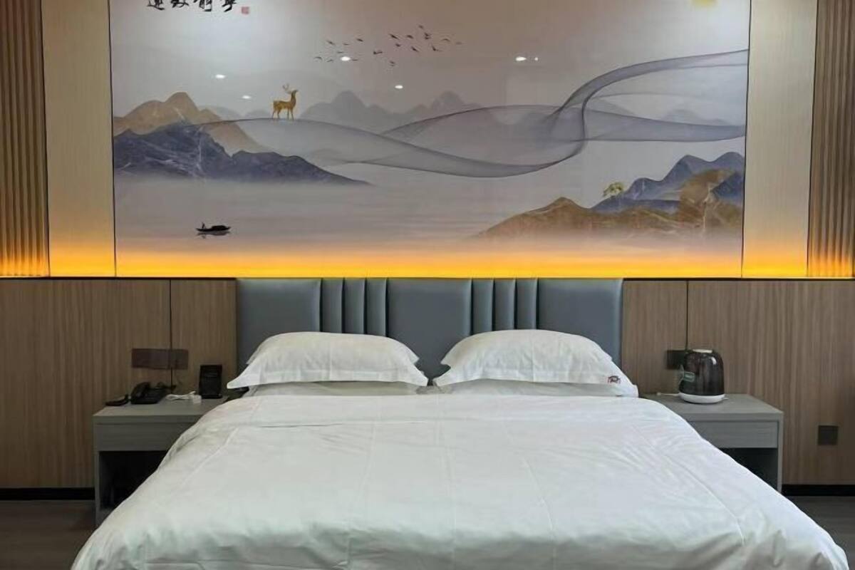 Una habitación de hotel con cama, mesita de noche, una obra de arte en la pared que representa un paisaje e iluminación en el techo.
