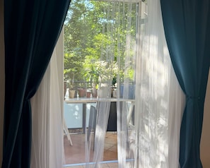 A view through a window with sheer curtains, revealing a balcony with potted plants and outdoor furniture.