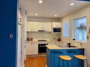 A compact kitchen with white cabinets, a microwave, a stove, and a sink. There are two wooden stools and a window with blinds.