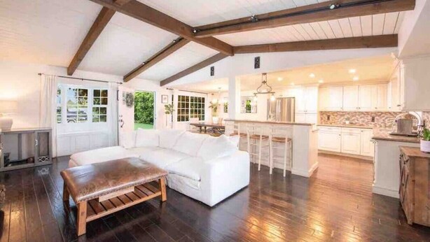 A spacious living area with a white sectional sofa, a wooden bench, and a kitchen with white cabinets and a stone backsplash. - Charming Ranch w/spa, 1/2 acre near Miramar Beach (Montecito)