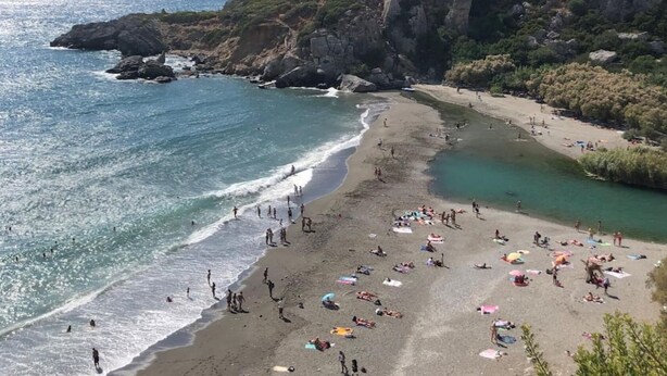 Ein Strand mit badenden und sonnenbäudelnden Personen, umgeben von Klippen und Grünpflanzen.