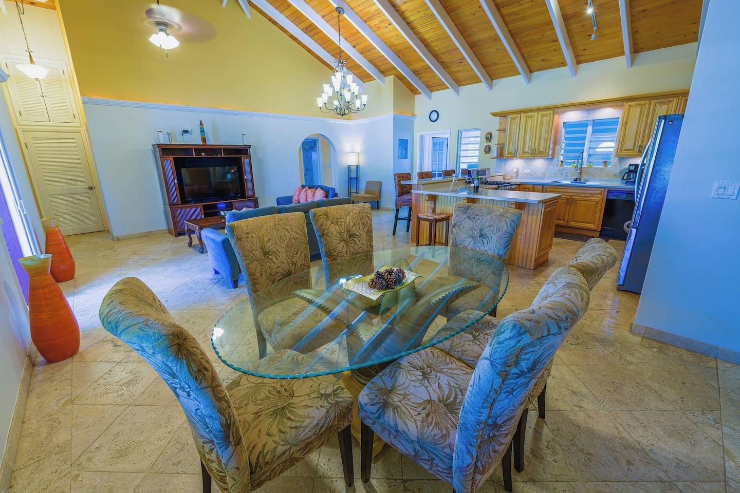 A spacious living area with a glass-top dining table surrounded by patterned chairs, a kitchen with wooden cabinets, and a ceiling with exposed beams.