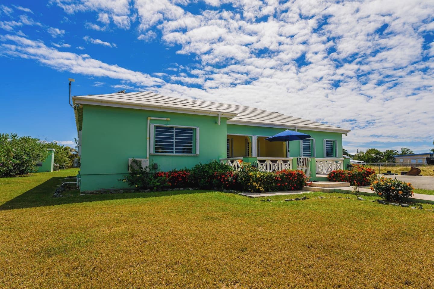 A single-story house with a green exterior, a front porch with a blue umbrella, and a small garden with red flowers.