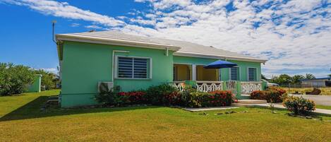 A single-story house with a green exterior, a front porch with a blue umbrella, and a small garden with red flowers.