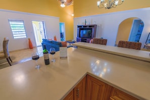 Una cocina con encimera, copas de vino y botellas. Hay un televisor sobre un soporte, un sofá azul y una silla de mimbre.