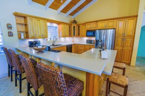 A kitchen with wooden cabinets, a stainless steel refrigerator, a microwave, and a large island with bar stools. - Perfect 4BDRM for families or groups! (Frederiksted)