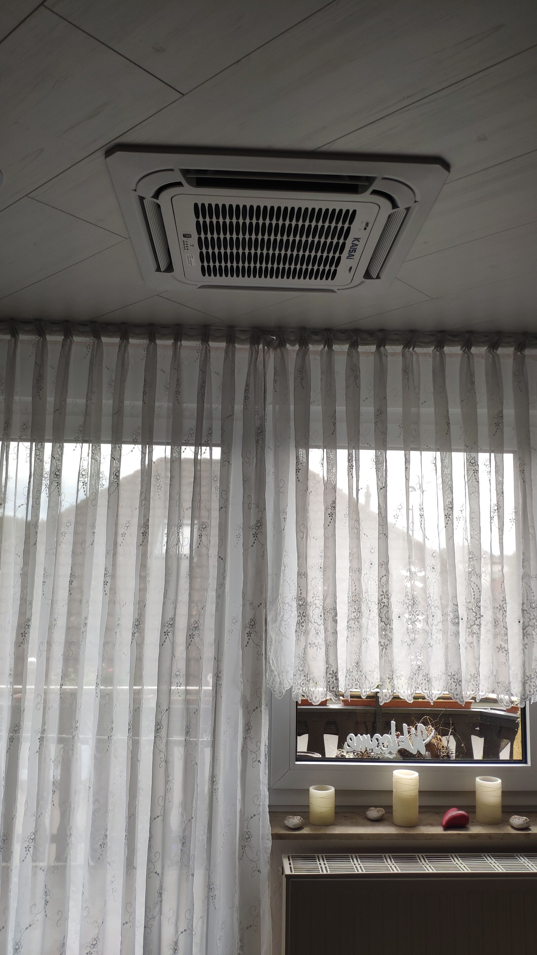A room with a ceiling vent, sheer curtains with floral patterns, a window sill with decorative items, and a vented air conditioning unit.