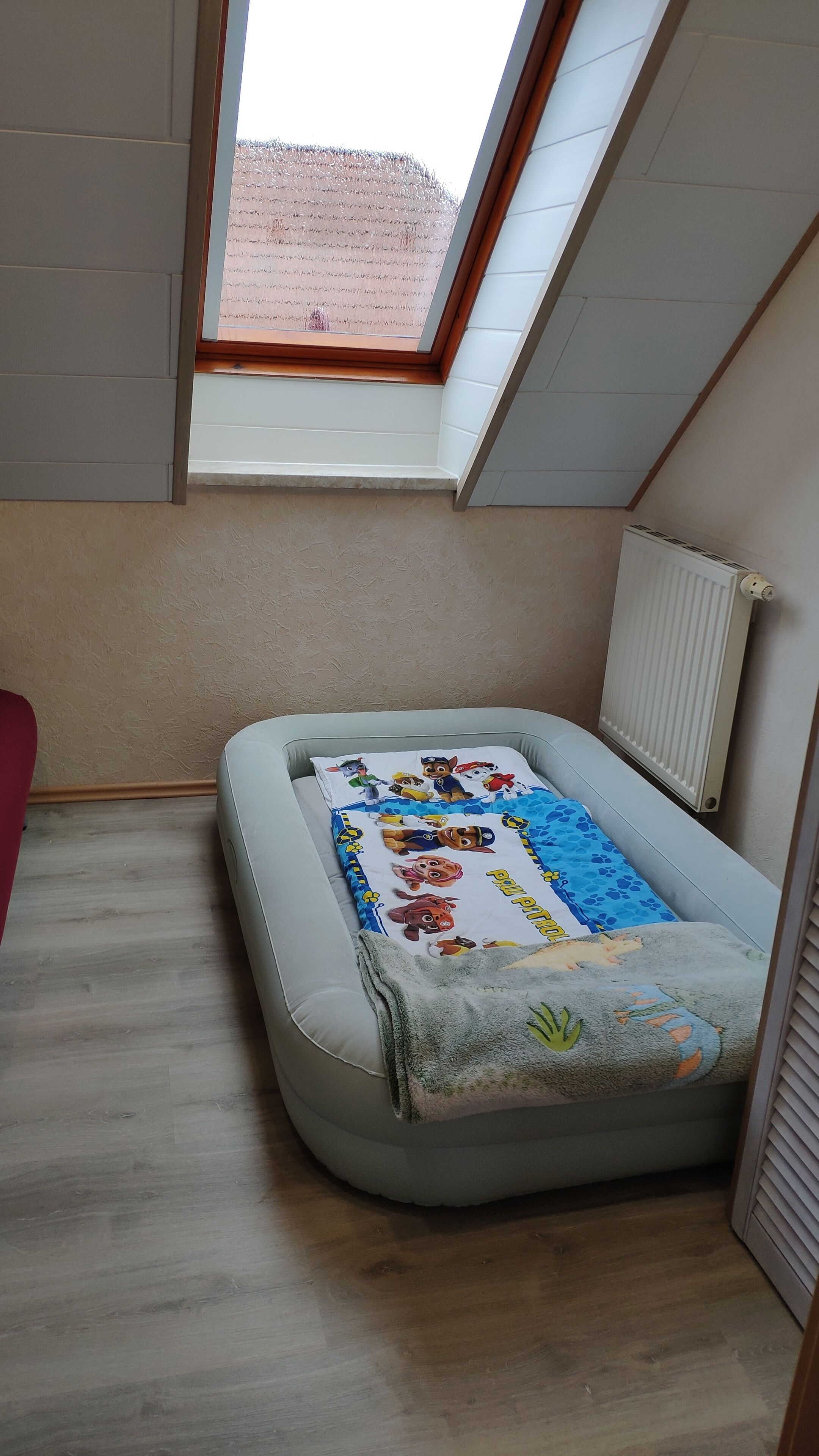 Ein kleiner Raum mit schrägem Dach, einem Fenster, einem Heizkörper und einem Bett mit bunter Decke.