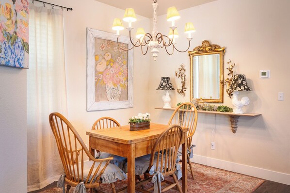 Un coin repas avec une table et des chaises en bois, un lustre, un miroir et des œuvres d’art florales accrochées aux murs.