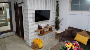 A living room with a TV mounted on a white brick wall, a brown sofa with a cushion, a wooden coffee table with a basket and flowers, and a patterned rug.