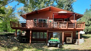 A two-story wooden cabin with a balcony, a covered patio, and a green vehicle parked in front. - Poskin Lake Getaway Cabin Lakefront access and beautiful sunrise (Almena)