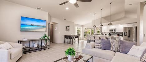 A spacious living area with a ceiling fan, a large TV, and a kitchen in the background.