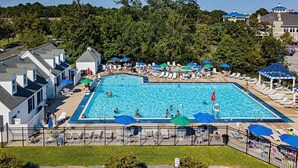 Una grande piscina rettangolare circondata da lettini e ombrelloni, con una recinzione e un edificio sullo sfondo.