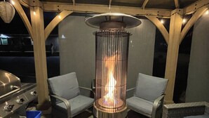 Une terrasse extérieure dotée d’un foyer central, de deux fauteuils gris rembourrés, d’un barbecue et d’une poubelle bleue.