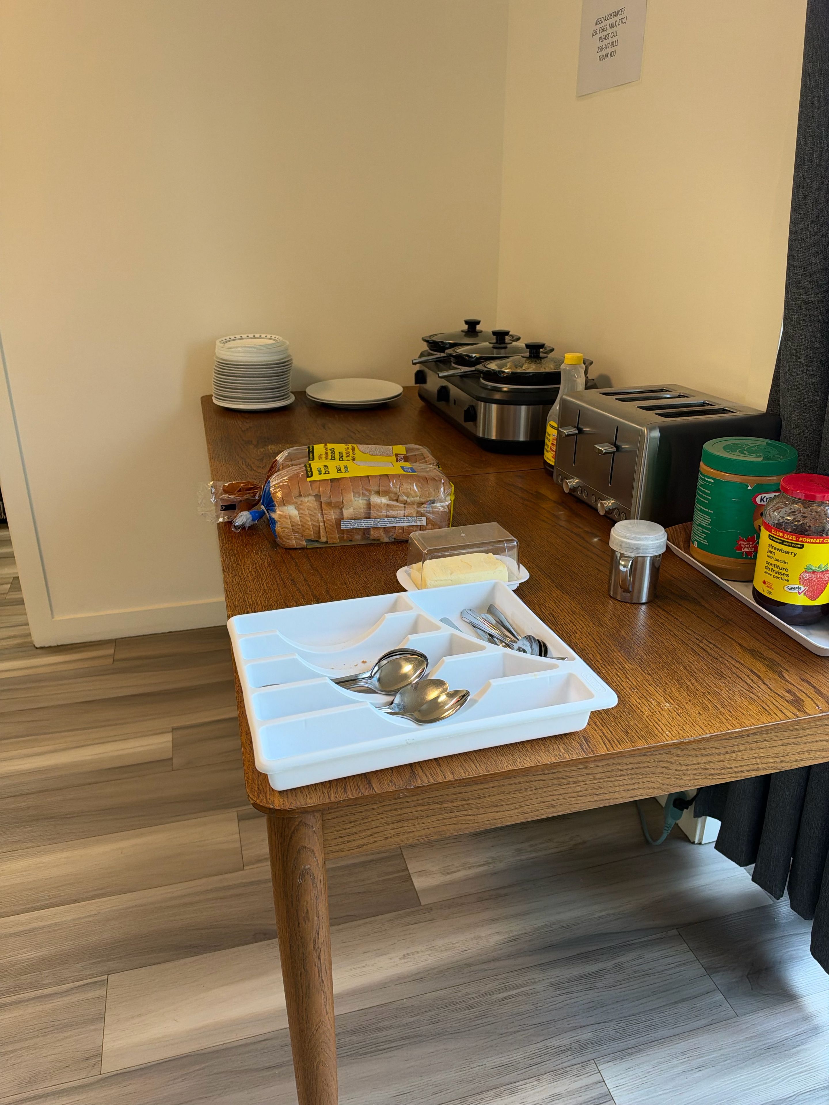 A kitchen area with a wooden table, a toaster, a kettle, a bread loaf, a butter dish, a jar of jam, and a white tray with utensils.