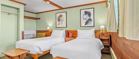 A double room with two beds, a wooden bench, a small table, and a nightstand with a lamp. There are framed artworks on the wall.