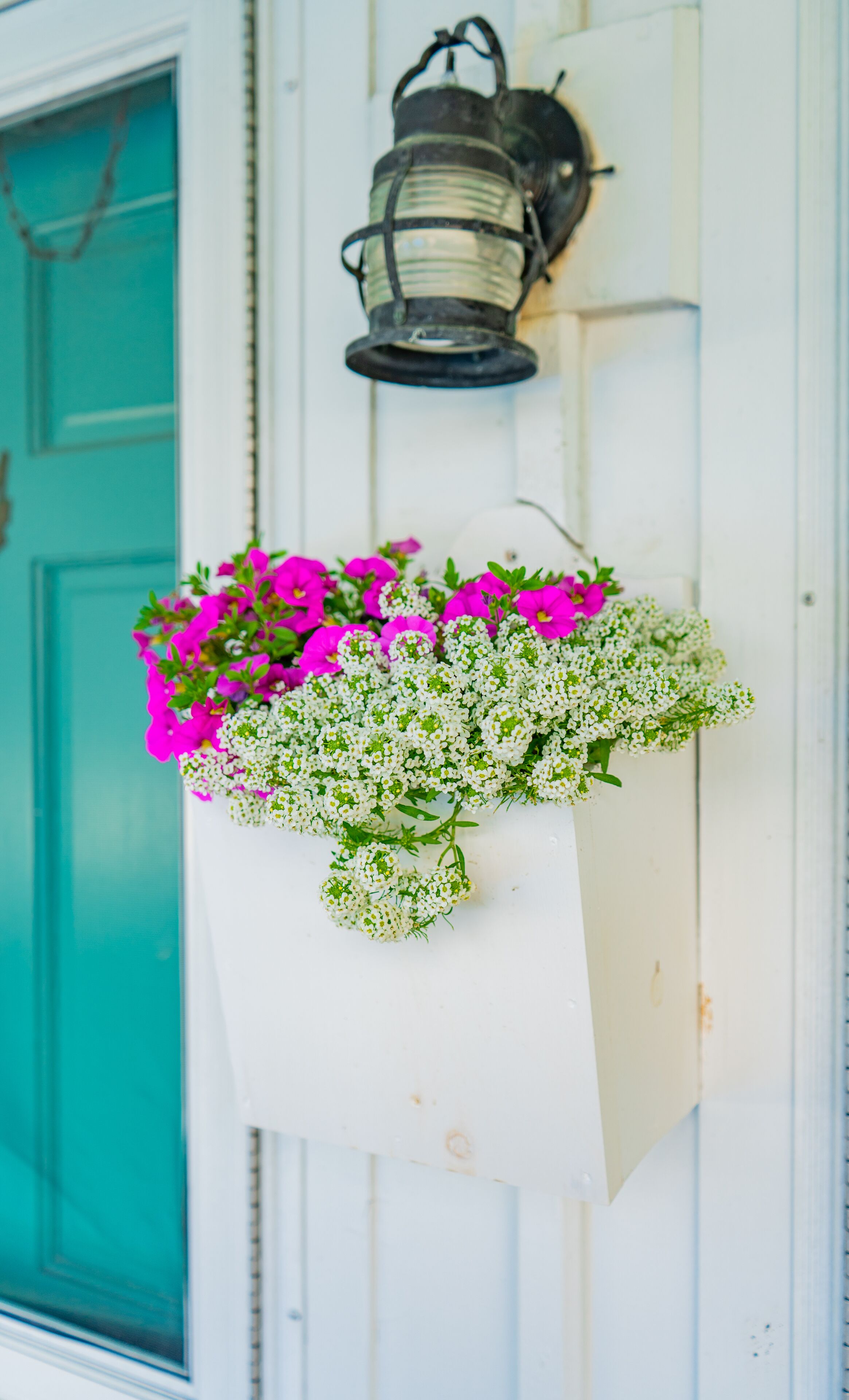 A wall-mounted flower basket with pink and white flowers, a teal door, and a black lantern.