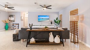 Un salon moderne avec un canapé gris, une table basse en bois et deux vases blancs. On y trouve également des ventilateurs de plafond, un téléviseur et une grande fenêtre avec des stores.