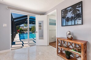 A modern interior with a glass door leading to a pool, a wooden console table, and a framed picture of palm trees.