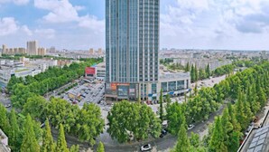 A high-rise building with a modern glass facade, surrounded by a parking lot and trees. - Thank You Intercity Hotel (Junan Leshang Building) (Junan County)