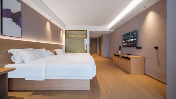 A modern hotel room with a large bed, a wooden bedside table, a built-in TV, and a small seating area. - Thank You Intercity Hotel (Junan Leshang Building) (Junan County)