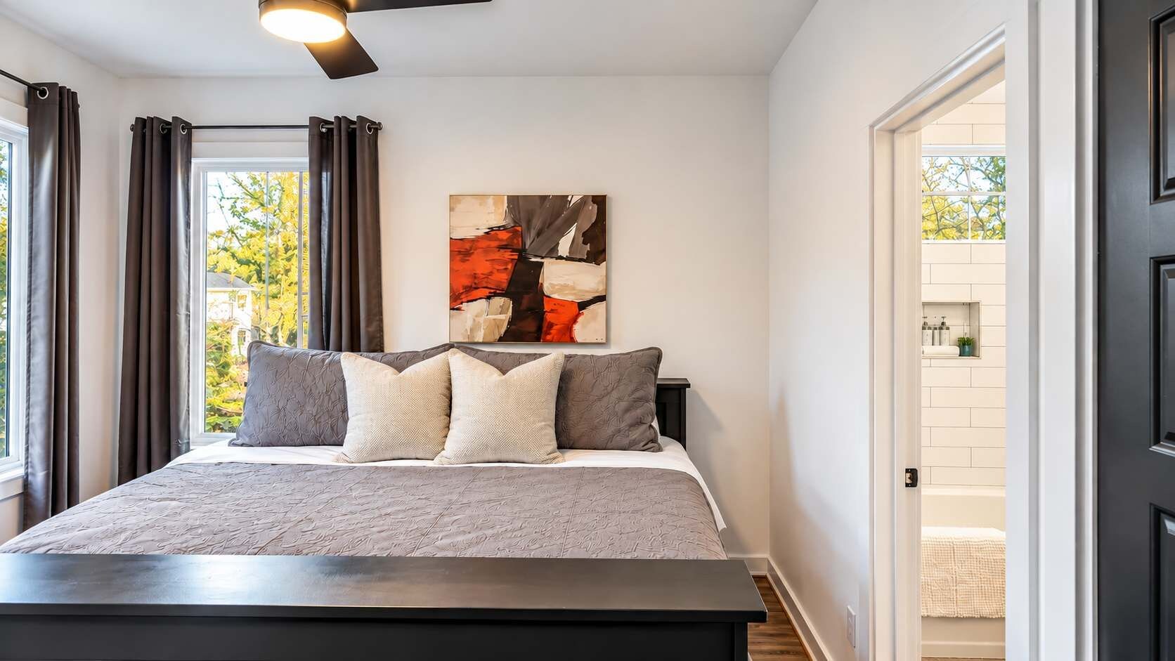 Un dormitorio con cama, ventilador de techo, ventana con cortinas y un cuadro en la pared.