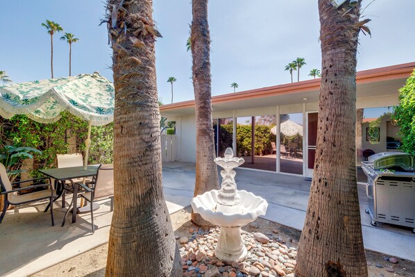A patio area with a stone fountain, palm trees, outdoor seating, and a building with large windows in the background.