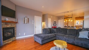 A living room with a sectional sofa, a fireplace, a television, and a dining area in the background. - Chalet Au petit Edouard (Mount Edouard) (L'Anse-Saint-Jean)