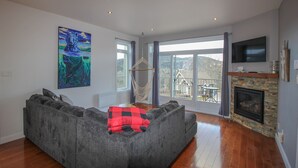 A modern living room with a grey sectional sofa, a fireplace, and a large window offering a view of the outdoors. - Chalet Au petit Edouard (Mount Edouard) (L'Anse-Saint-Jean)