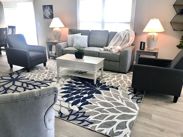 A living room with a grey sofa, two armchairs, a white coffee table, and a patterned rug. There are two lamps on side tables, and a plant on the coffee table.