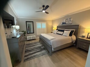 A bedroom with a bed, bedside tables, a nightstand, a ceiling fan, a mirror, and a window with blinds.