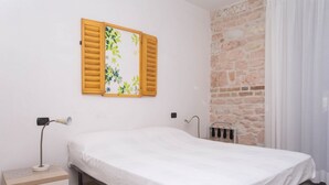 A single bed with a white mattress, a nightstand with a lamp, a radiator, and a window with wooden shutters. - A03 - Massignano, two room apartment (Ancona)
