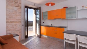 A modern kitchen with orange cabinets, a dining area with white chairs, and a stone wall. - A03 - Massignano, two room apartment (Ancona)