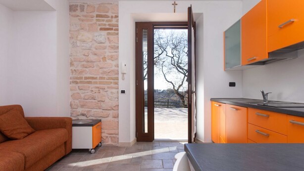 A modern kitchen with orange cabinets, a stone wall, and a view through an open door. - A03 - Massignano, two room apartment (Ancona)