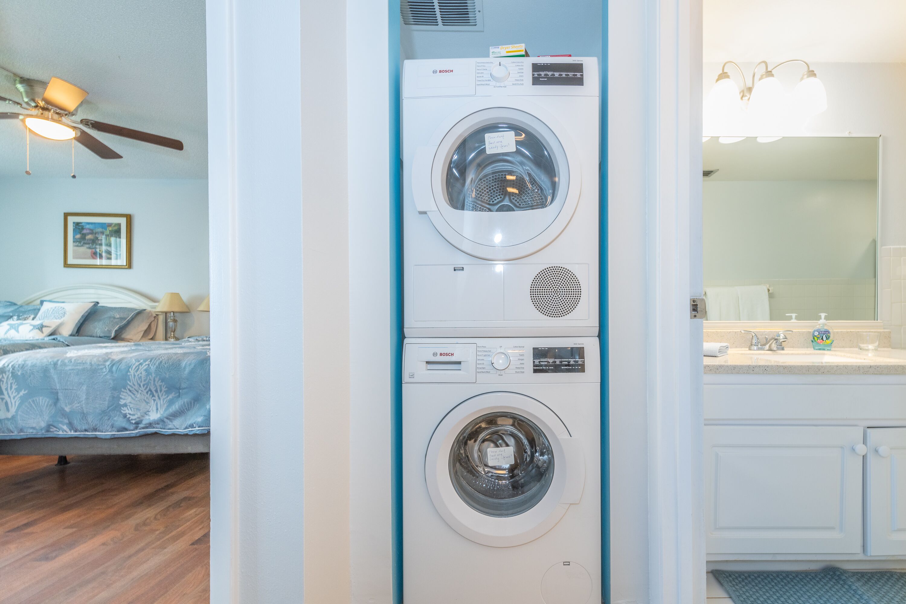 A laundry area with two stacked washing machines, a bed with a blue patterned bedspread, a ceiling fan, and a bathroom with a sink and mirror.