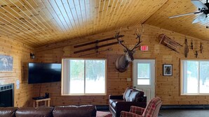 A log cabin interior with a fireplace, a television, a large window, a brown leather sofa, a plaid armchair, and a mounted deer head.