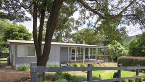 A small, single-story house with a covered porch, surrounded by a wooden fence and various shrubs.