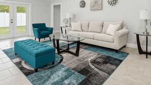 A modern living room with a white sofa, teal armchair, and ottoman, a glass coffee table, and a patterned rug. - Entire House close to everything. (Seffner)