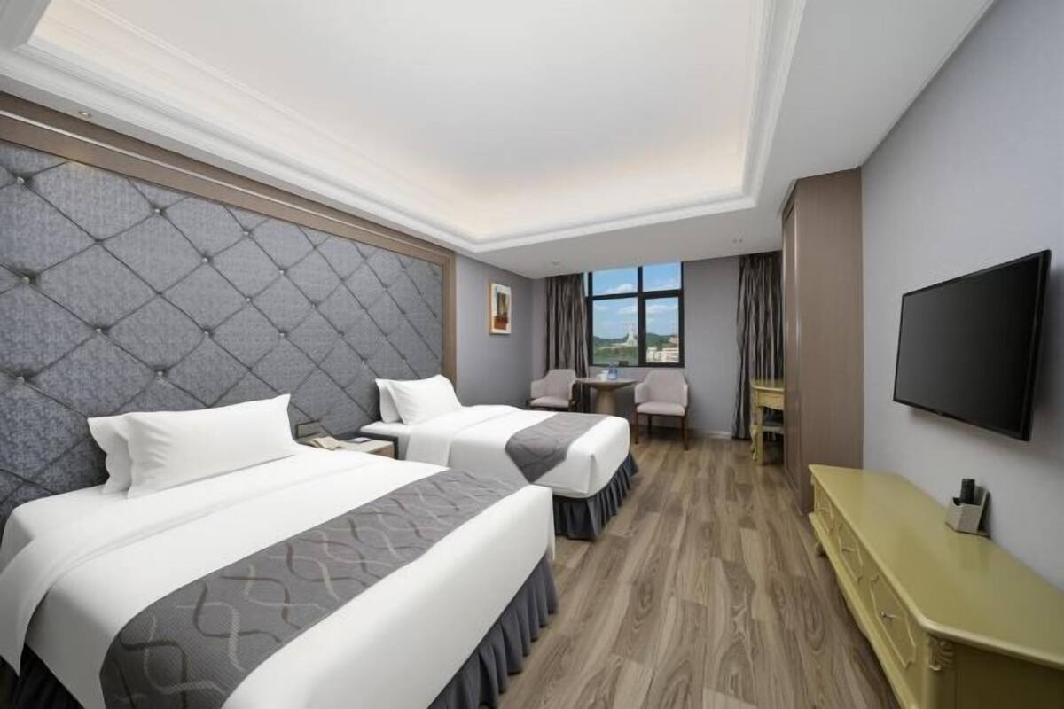 A hotel room with two beds, a TV, a small table, and a view of the outdoors.