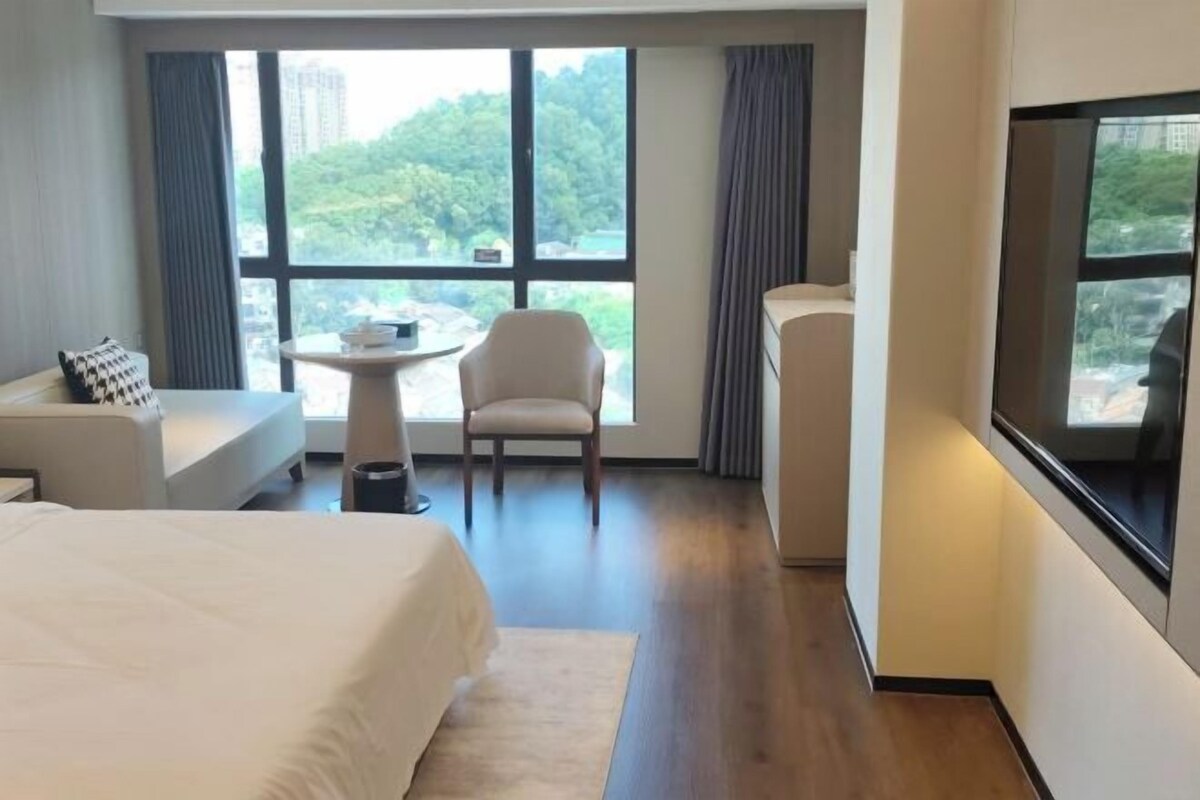 Habitación de hotel con una cama grande, una mesita con una silla, televisor montado en la pared y ventana con vistas a zonas verdes.