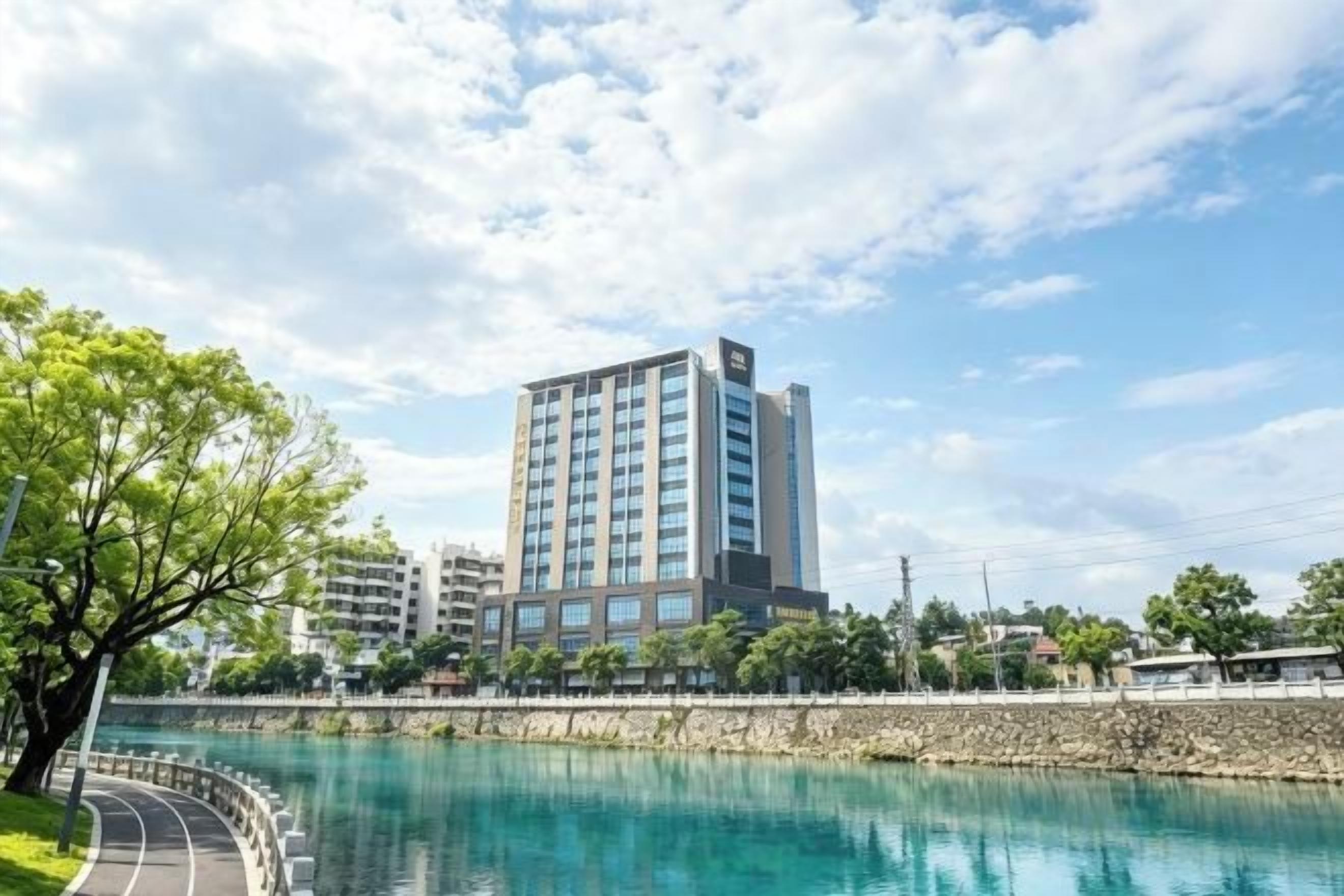A modern hotel with a view of a river, surrounded by greenery and other buildings.