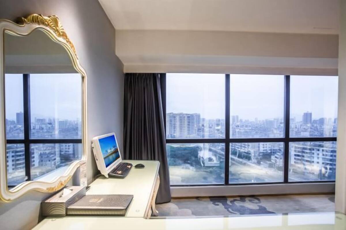 A modern hotel room with a large window, a desk with a computer, a mirror, and a view of the cityscape.