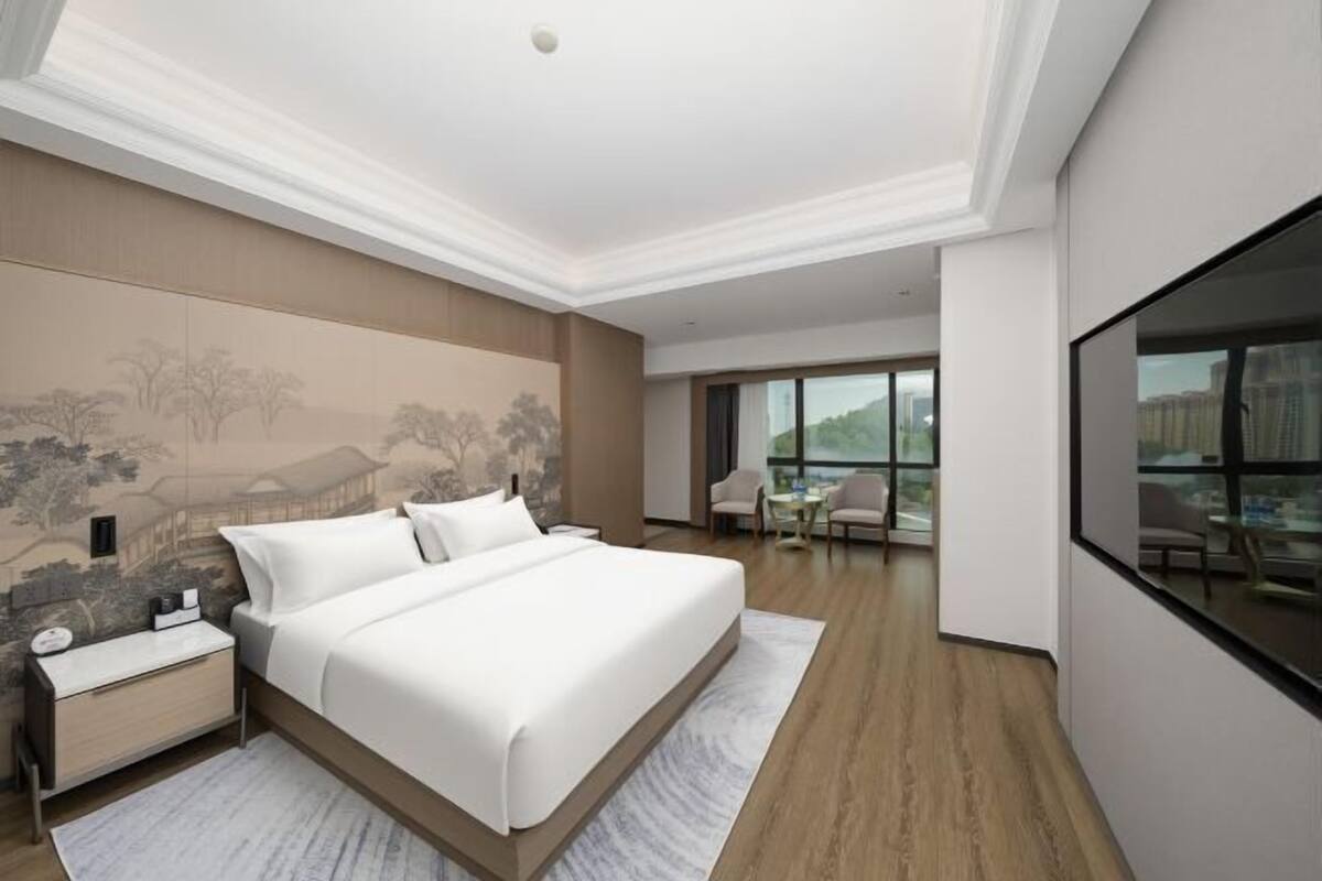 A modern hotel room with a large bed, a nightstand, a desk, and a view of the city through large windows.