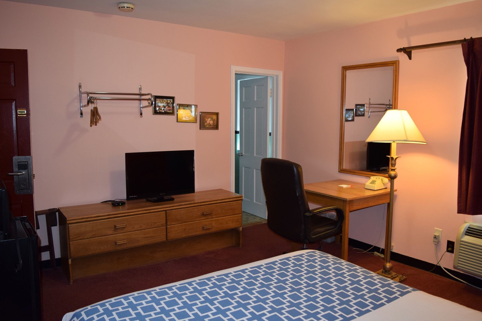 Hotellihuone, jossa on sänky, kirjoituspöytä lampun kanssa, televisio, tuoli, lipasto ja ovi.
