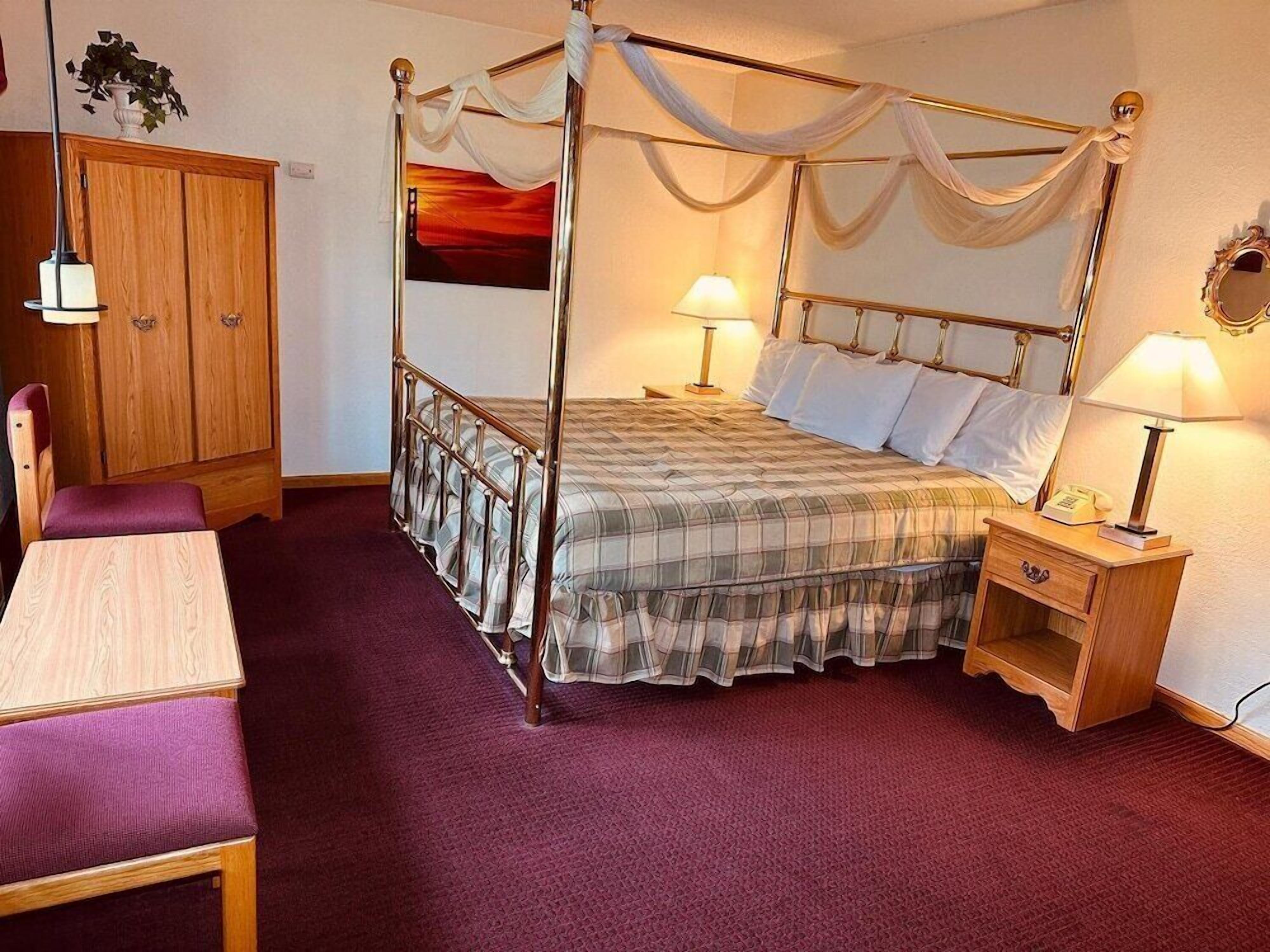 A four-poster bed with a plaid bedspread, two bedside lamps, a wooden nightstand, a wooden wardrobe, and a small table with a purple cushion.
