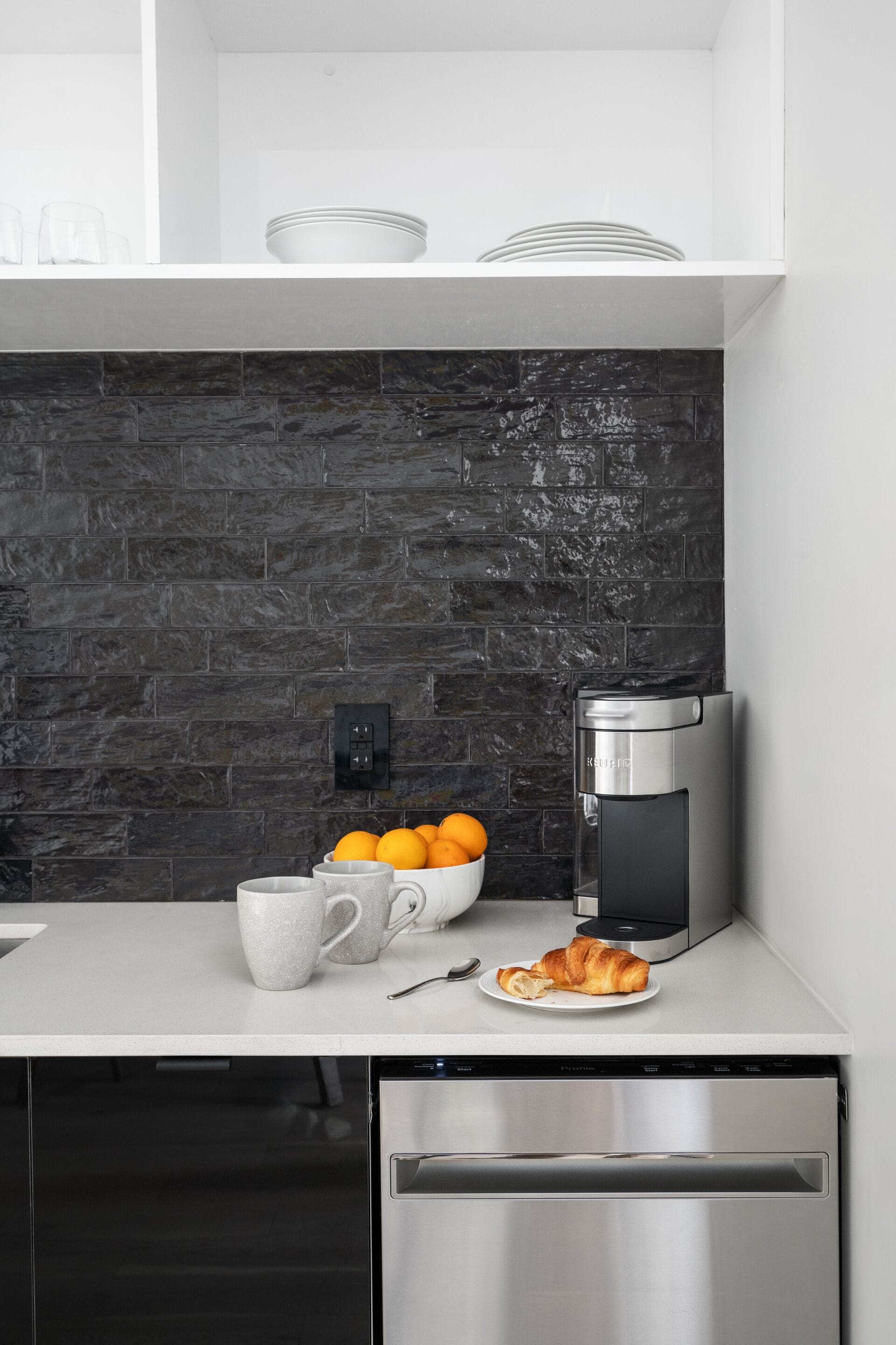 A modern kitchen with a dark brick backsplash, a coffee maker, a bowl of oranges, and a plate with a croissant.