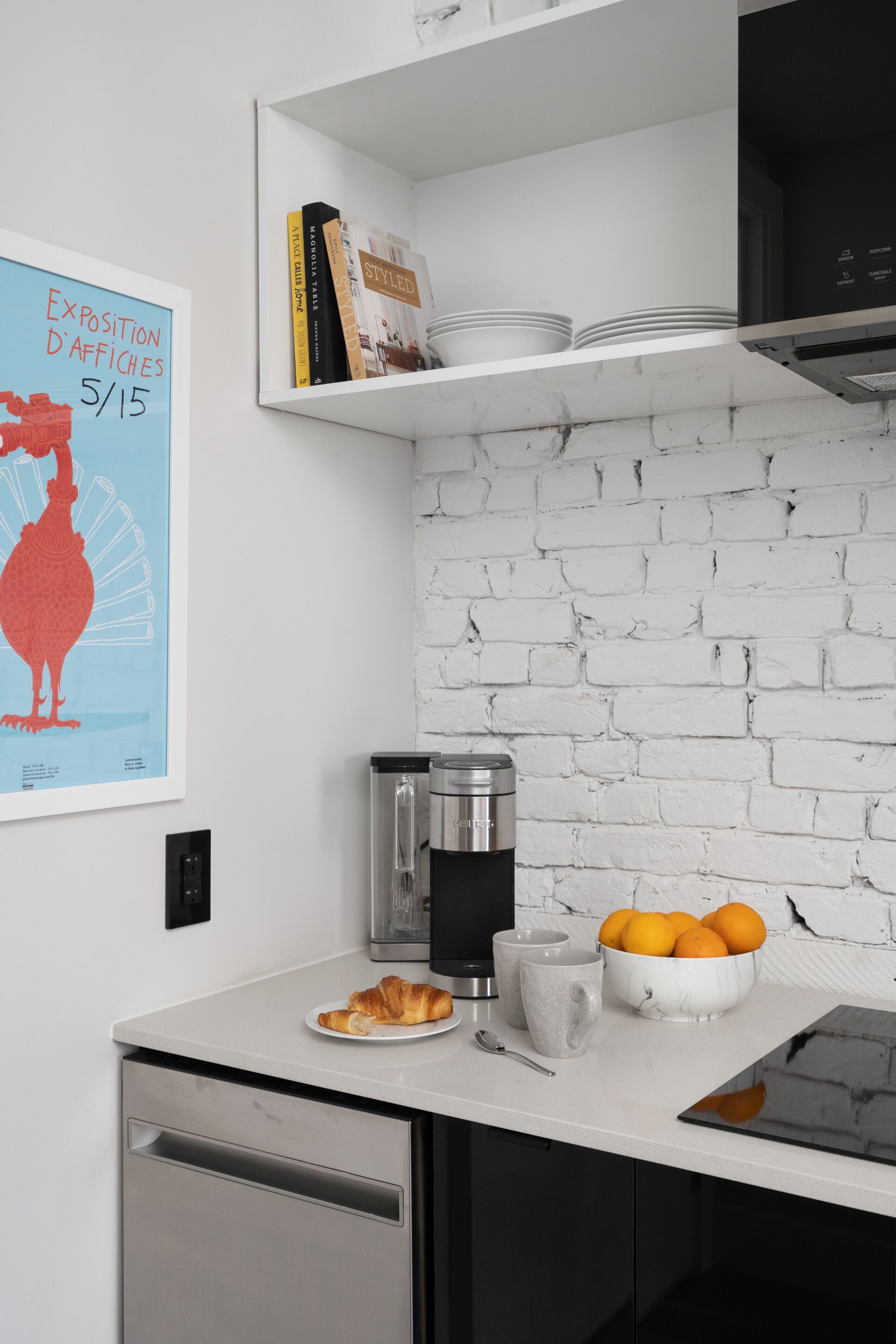 A modern kitchen with white brick backsplash, a coffee machine, a bowl of oranges, and a croissant on the counter.