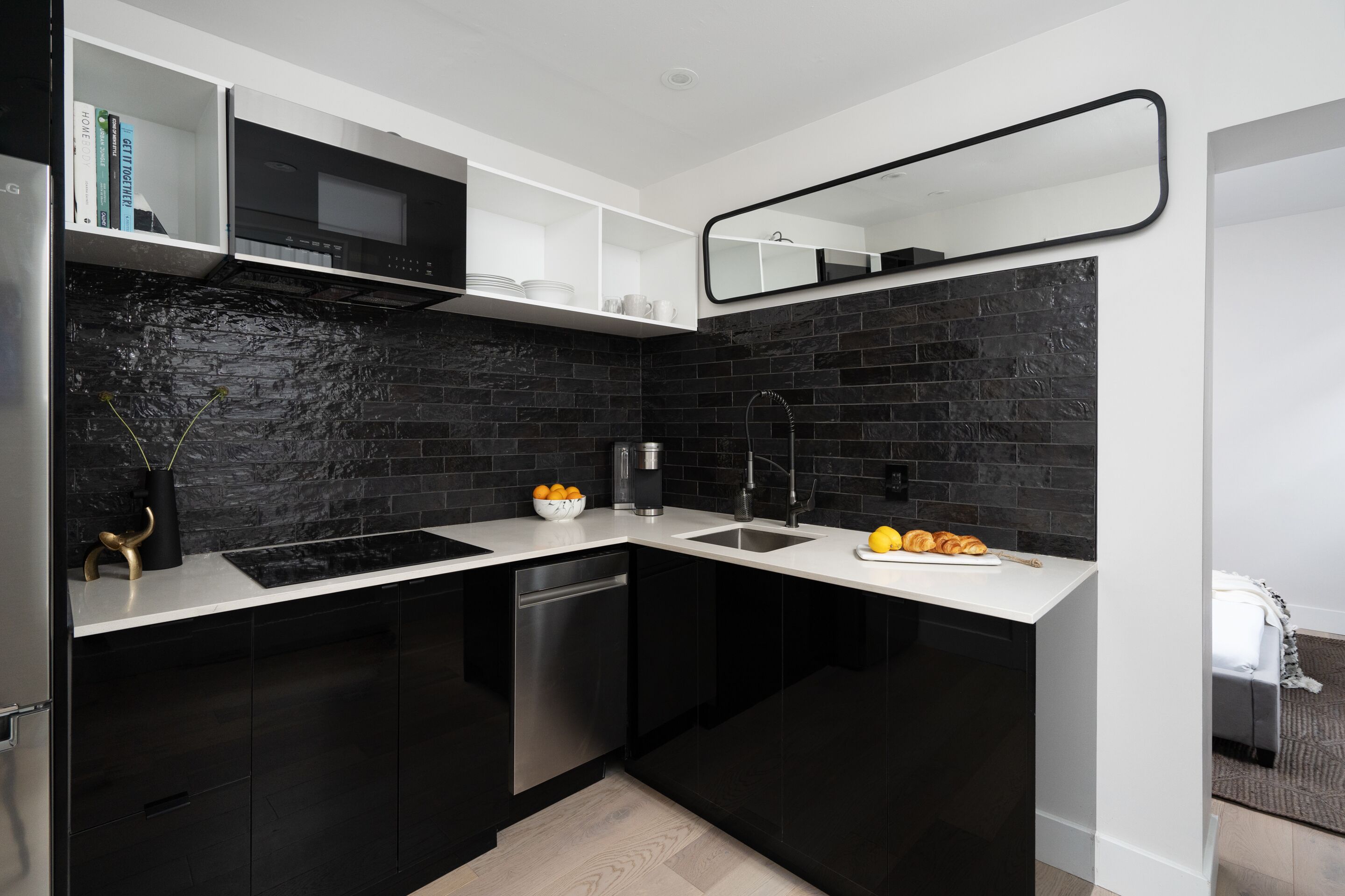 Una cocina moderna con gabinetes negros, encimera blanca, microondas integrado y un espejo grande sobre el fregadero.