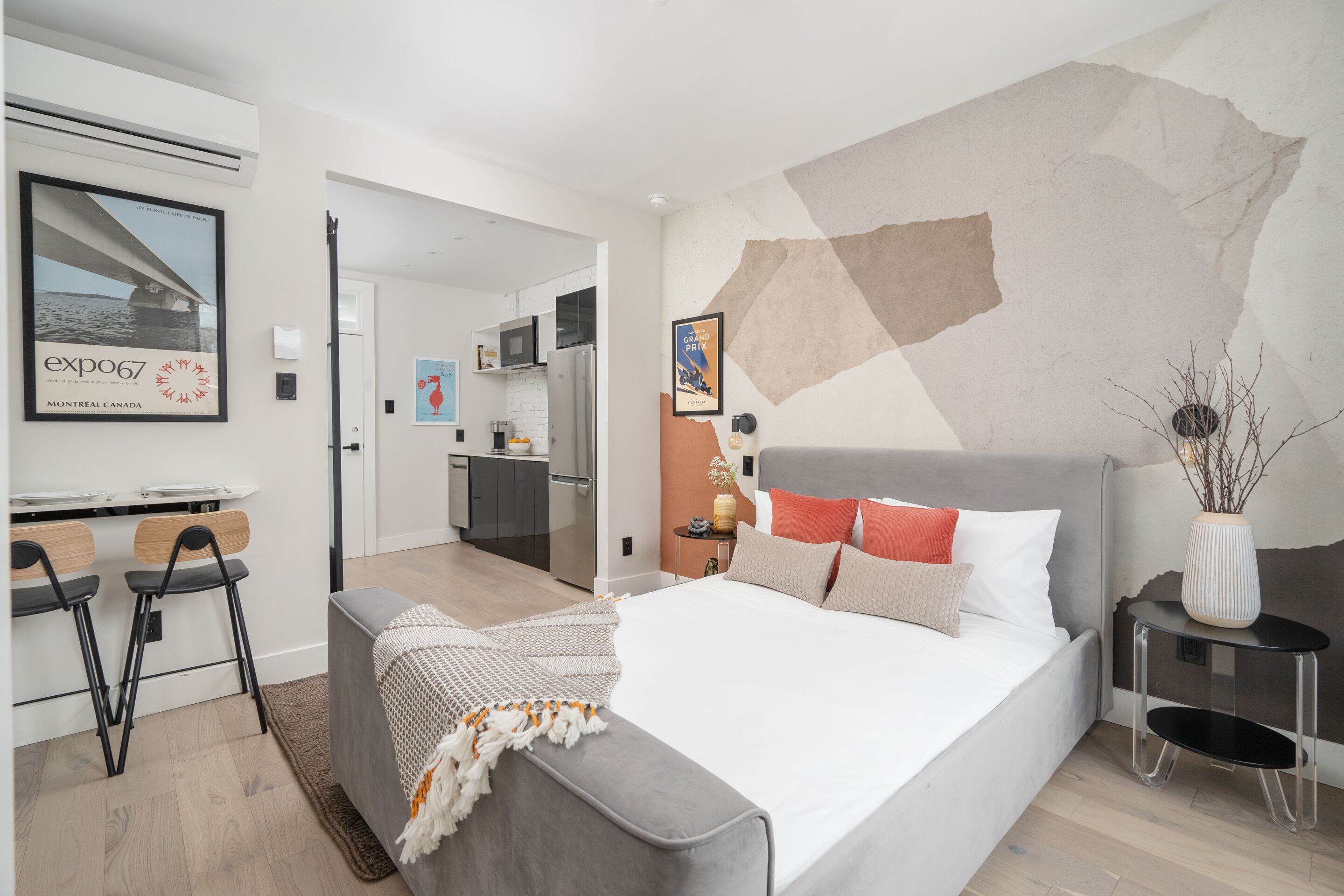 A modern hotel room with a large bed, a desk with two chairs, a kitchenette, and a view of the city.
