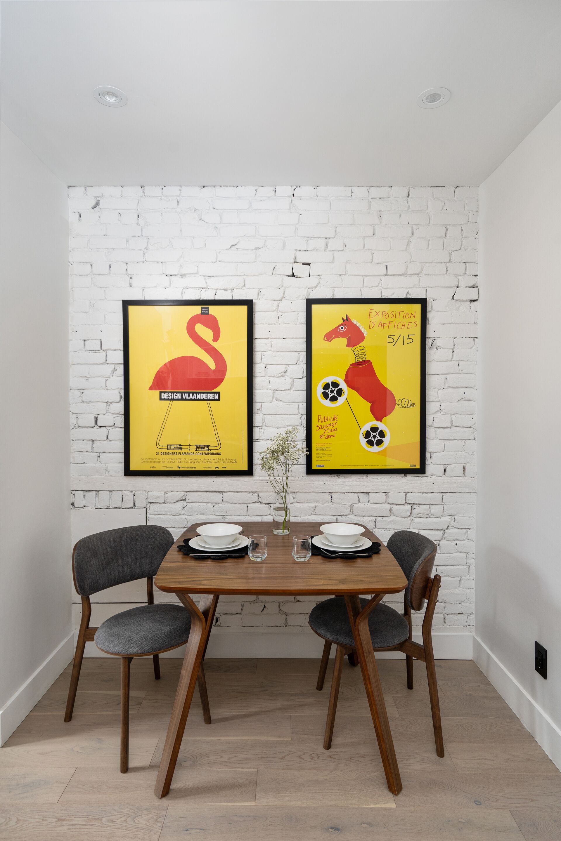 Un área de comedor con pared de ladrillo, dos carteles enmarcados, una mesa de madera, cuatro sillas grises y un pequeño florero con flores.